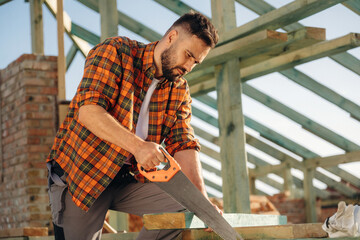 Focused, holding saw and cutting the plank. Construction worker is on the roof of a house