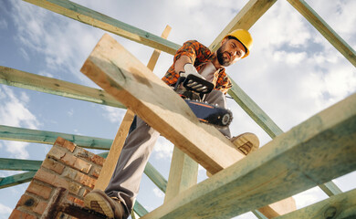 View from below, cutting plank by chainsaw. Construction worker is on the roof of a house