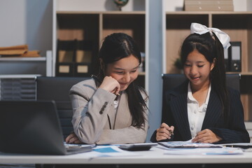 Fototapeta premium Two young Asian business woman talk, consult, discuss working with new startup project idea presentation analyze plan marketing and investment in the office.