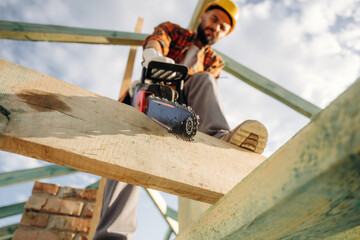 Serious facial expression, cutting the plank. Construction worker is on the roof of a house