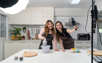 Two asian women in an apron, smiling and cooking in a kitchen with a smartphone on a selfie stick and lighting equipment for video and photo shoot, recording vlog video for social blogger.
