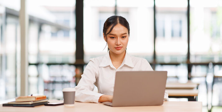 Smiling asian female marketing manager, professional it specialist working, browsing at laptop computer sitting at desk in modern office. Cheerful young woman employee work for business