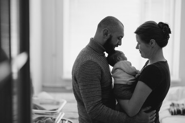 A couple lovingly holds their newborn baby in a cozy indoor setting. The mother gazes at the baby...
