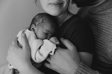 A couple lovingly holds their newborn baby in a cozy indoor setting. The mother gazes at the baby...