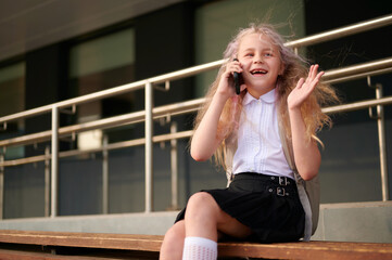 cheerful girl using smartphone while sitting outdoors school building, enjoying conversation. young student with backpack displaying joy, connectivity, technology in urban environment