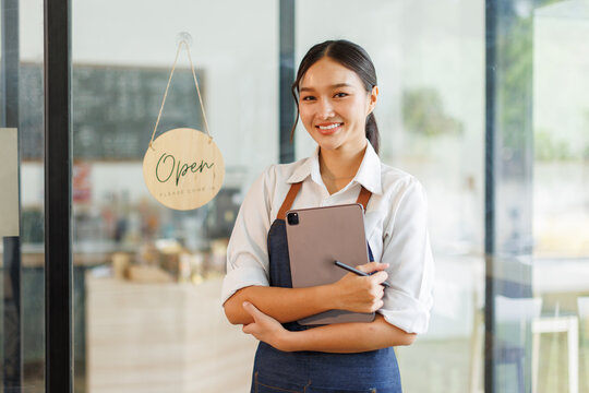 Portrait of happy asian woman standing and holding digital tablet at open sign of her store.  Successful small business owner in casual wearing blue apron standing at entrance cafe.
