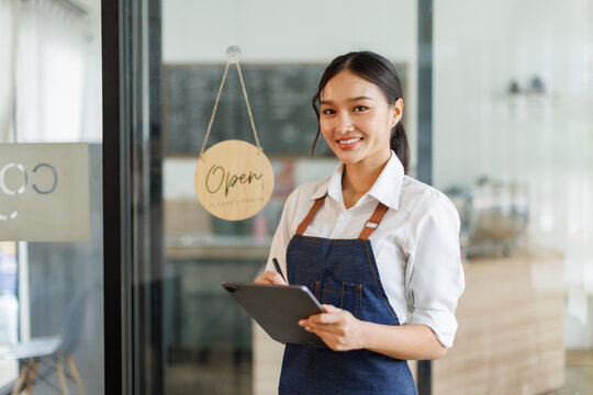 Portrait of happy asian woman standing and holding digital tablet at open sign of her store.  Successful small business owner in casual wearing blue apron standing at entrance cafe.
 - Powered by Adobe