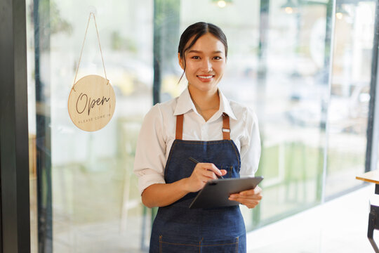 Portrait of happy asian woman standing and holding digital tablet at open sign of her store.  Successful small business owner in casual wearing blue apron standing at entrance cafe.
