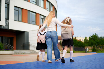 mother walking children to school, girls in uniforms with backpacks. building exterior suggests...