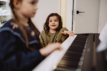 Two young girls playing a piano together, one smiling and looking at the other. The setting is...