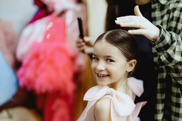 A young girl with a big smile is having her hair styled by an adult. She is wearing a pink dress...