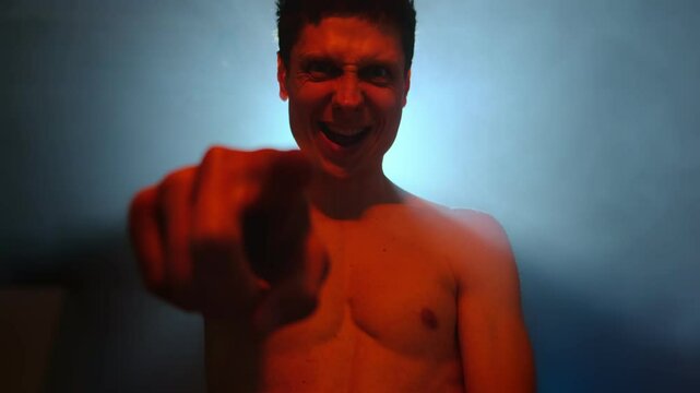 Man laughing hysterically and pointing finger, in red and blue smoke-lit scene of madness and emotional chaos.