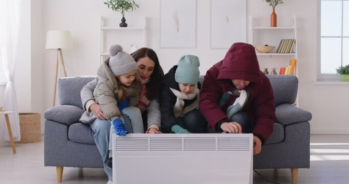 Frozen family with children dressed in winter outerwear struggles with the freezing weather inside home in living room, seeking warmth near a heater. Cold conditions and the need for warmth.