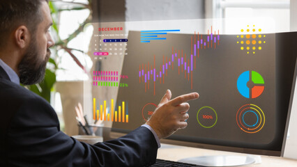 Professional in suit points to colorful digital statistics and charts, including calendar and pie graphs, on transparent screen in modern office. Concept of business planning, workplace analytics