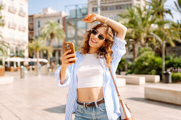 A joyful young woman in sunglasses and holding a phone in her hands walks through sunny streets. A...