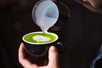 A barista carefully pours steamed milk into a cup of matcha latte to create latte art. The heart...