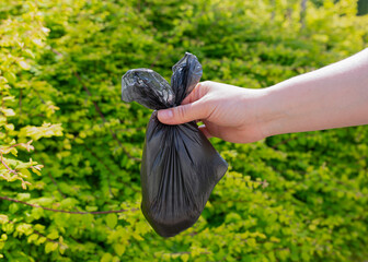 Close-up of a human hand holding a small black plastic bag with dog poop, against a green park...