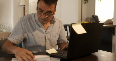 A mature, bespectacled man with a worried look on his face reviews bills, using a laptop as a calculator, in his living room, - Powered by Adobe