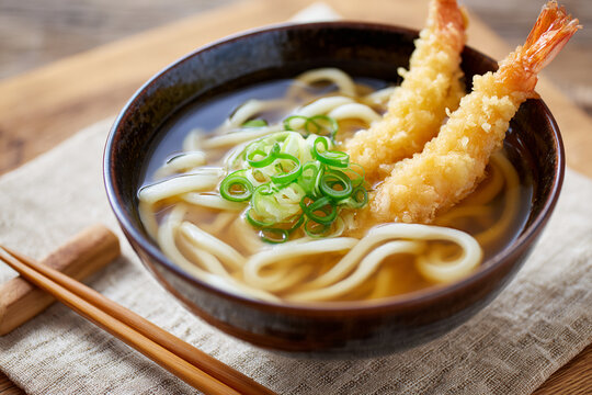 Bowl of udon noodles in clear broth with floating scallions and two tempura shrimp leaning over the edge, wooden chopsticks on linen to the left