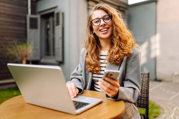 A young woman sits at a table on the veranda in a cafe with a laptop and a phone in her hands. A...