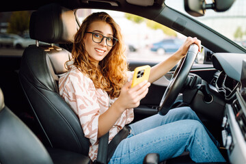 A young woman with curly hair driving a car and holding a phone in her hands on a sunny day. Woman...