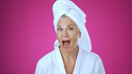 A happy mature woman in a spa setting looks at her reflection in a handheld mirror, experiencing joy and relaxation in a serene, pink background.
