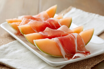 Thin slices of cantaloupe melon wrapped with prosciutto arranged asymmetrically on white ceramic platter