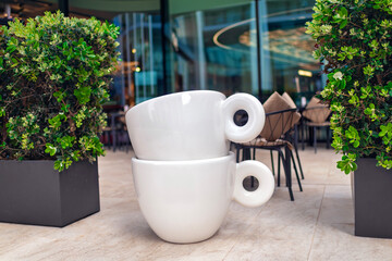  Giant white coffee cups placed outdoors among green plants at a street cafe, with stylish chairs...