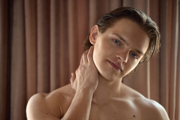 A young man gently touches his neck, posing relaxedly against a soft brown curtain backdrop. Concept of tranquility and self-care in a serene environment.