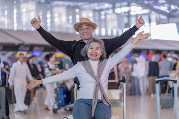 happy asian senior woman sitting on luggage that put on trolley,raising hands up,her husband standing behind in airport,excited and ready to travel