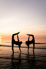 Silhouette of young women standing in gymnastic poses in the water at sunset. Beautiful dancers demonstrate acrobatic skills against a bright sky. Concept of sport, nature and relaxation.