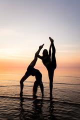 Silhouette of young women standing in gymnastic poses in the water at sunset. Beautiful dancers demonstrate acrobatic skills against a bright sky. Concept of sport, nature and relaxation.