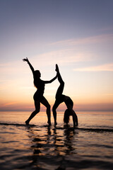 Silhouette of young women standing in gymnastic poses in the water at sunset. Beautiful dancers demonstrate acrobatic skills against a bright sky. Concept of sport, nature and relaxation.