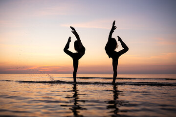 Silhouette of young women standing in gymnastic poses in the water at sunset. Beautiful dancers demonstrate acrobatic skills against a bright sky. Concept of sport, nature and relaxation.