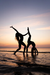 Silhouette of young women standing in gymnastic poses in the water at sunset. Beautiful dancers demonstrate acrobatic skills against a bright sky. Concept of sport, nature and relaxation.