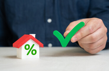 A man holds a check mark near his house. Mortgage approval at a favorable interest rate. Home...