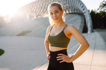 Portrait of a happy woman in sportswear standing in the rays of the sun on a sports ground. The...