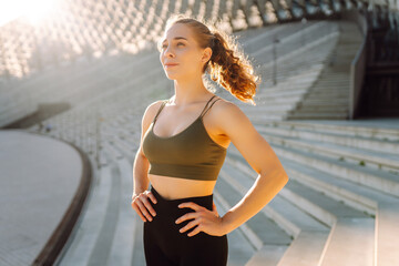 Portrait of a happy woman in sportswear standing in the rays of the sun on a sports ground. The athlete is warming up and enjoying the sunny weather outdoors. Sports concept. Active lifestyle.
