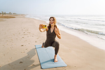 Young woman in sportswear doing yoga on a mat on the beach at sunrise. Athletic woman with wireless...