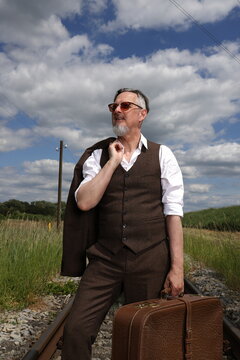 Mid-aged man with sunglasses walks with a brown leather suitcase and jacket on railway tracks to his next destination, tall grass and threatening cloud backdrop