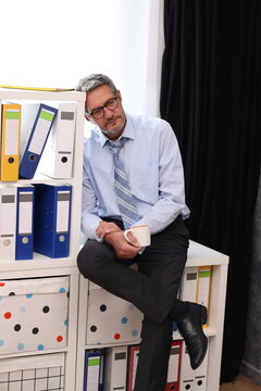 Office worker in shirt and tie sitting in office, leaning back, coffee cup in hand and looking exhausted