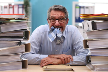 Office worker in shirt and tie sits in the office in front of stacks of files and eats his tie