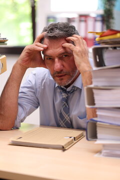 Office worker in shirt and tie sitting in the office in front of stacks of files and looks very annoyed and exhausted