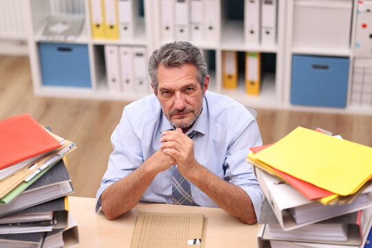 Office worker in shirt and tie sits in the office next to stacks of files and looks annoyed