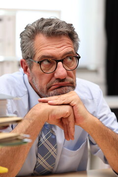 Office worker with glasses sits at the desk, supports his head with one arm and looks depressed and helpless to the side