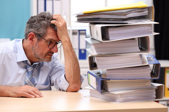 Office worker with glasses sits at the desk, supports his head with one arm and looks depressed to the side