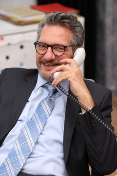 Office worker with glasses and suit sits at his desk, talks on the phone and is very satisfied with the conversation