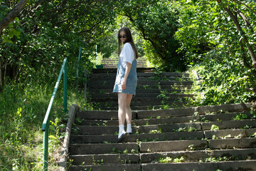 A young woman with long hair is descending stone stairs surrounded by greenery. She wears a simple...