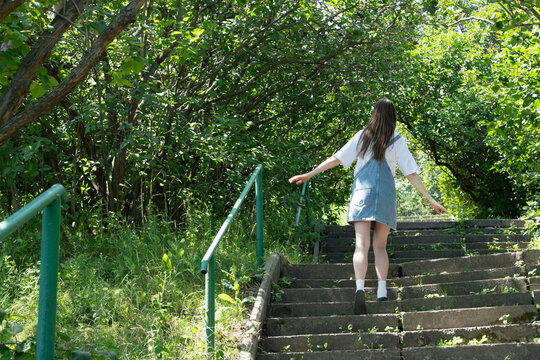A young woman in a casual outfit ascends a set of stone stairs surrounded by vibrant greenery. Sunlight filters through the trees, creating a serene atmosphere