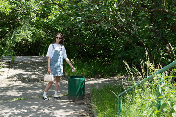 A young woman dressed in a denim dress and sunglasses strolls through a lush park. She stands near a green trash can, enjoying the sunny weather amid greenery